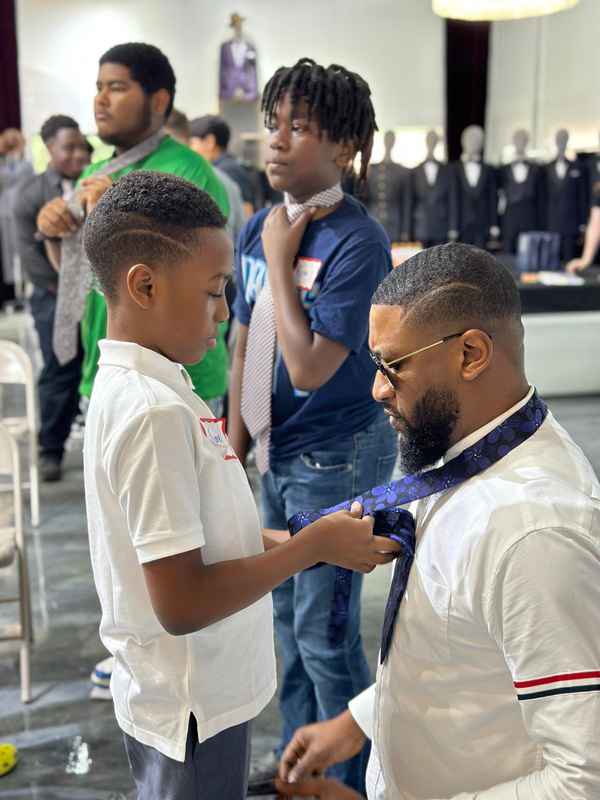 A young boy practicing tying a tie with help from a volunteer mentor