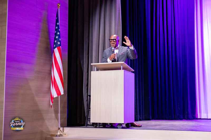 Guest speaker addressing the audience from a podium beside an American flag at the Miss Forney Scholarship Pageant
